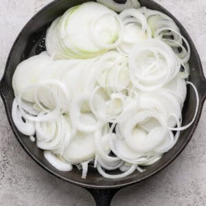 A cast iron skillet filled with raw, sliced white onions, arranged in overlapping rings on a light textured surface.
