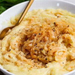 A bowl of mashed potatoes topped with caramelized onions and cracked black pepper, with a gold spoon on the side.