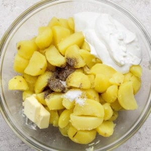 A glass bowl containing boiled, sliced potatoes, a pat of butter, a dollop of sour cream, salt, and black pepper on a light surface.