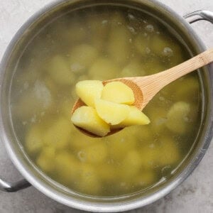 A pot of boiling water with peeled potato pieces, with several pieces being lifted out on a wooden spoon.