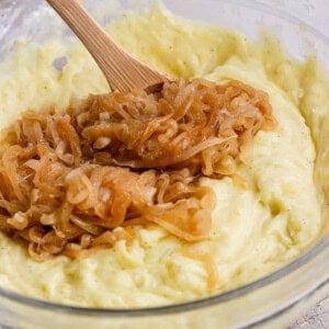 A glass bowl of mashed potatoes topped with caramelized onions, being mixed with a wooden spoon.