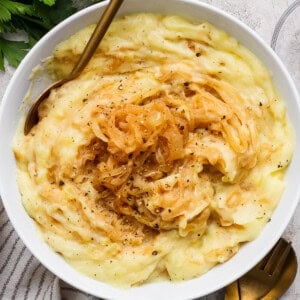 A bowl of mashed potatoes topped with caramelized onions and black pepper, with a gold fork and spoon beside the bowl on a light textured surface.