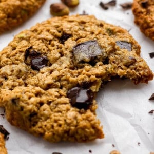 A close-up of an oatmeal chocolate chip cookie with a bite taken out, resting on white parchment paper with cookie crumbs around it.
