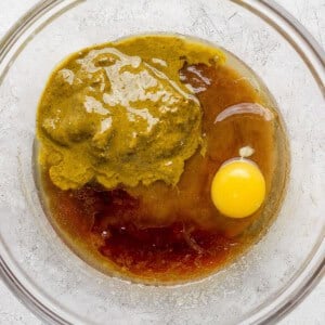 A clear glass bowl containing an egg, greenish nut butter, and brown liquid ingredients on a light textured surface.