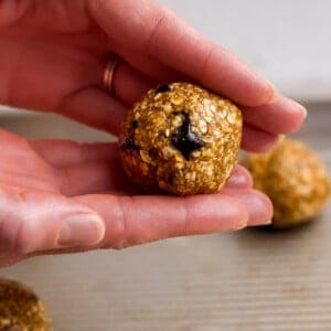 A hand holding a round, oat-based energy bite with a visible chocolate chip, above a metal baking tray with more energy bites in the background.