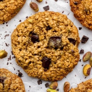 Oatmeal cookies with chocolate chunks and pistachios on parchment paper, surrounded by scattered chocolate pieces and pistachio nuts.