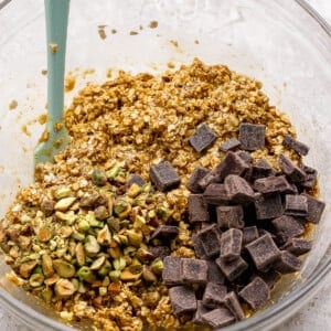 A glass bowl containing oat mixture with chopped pistachios and dark chocolate chunks, and a green spatula resting inside.