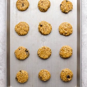 Twelve unbaked oatmeal cookies with chocolate chips are evenly spaced on a metal baking sheet, ready to be baked.