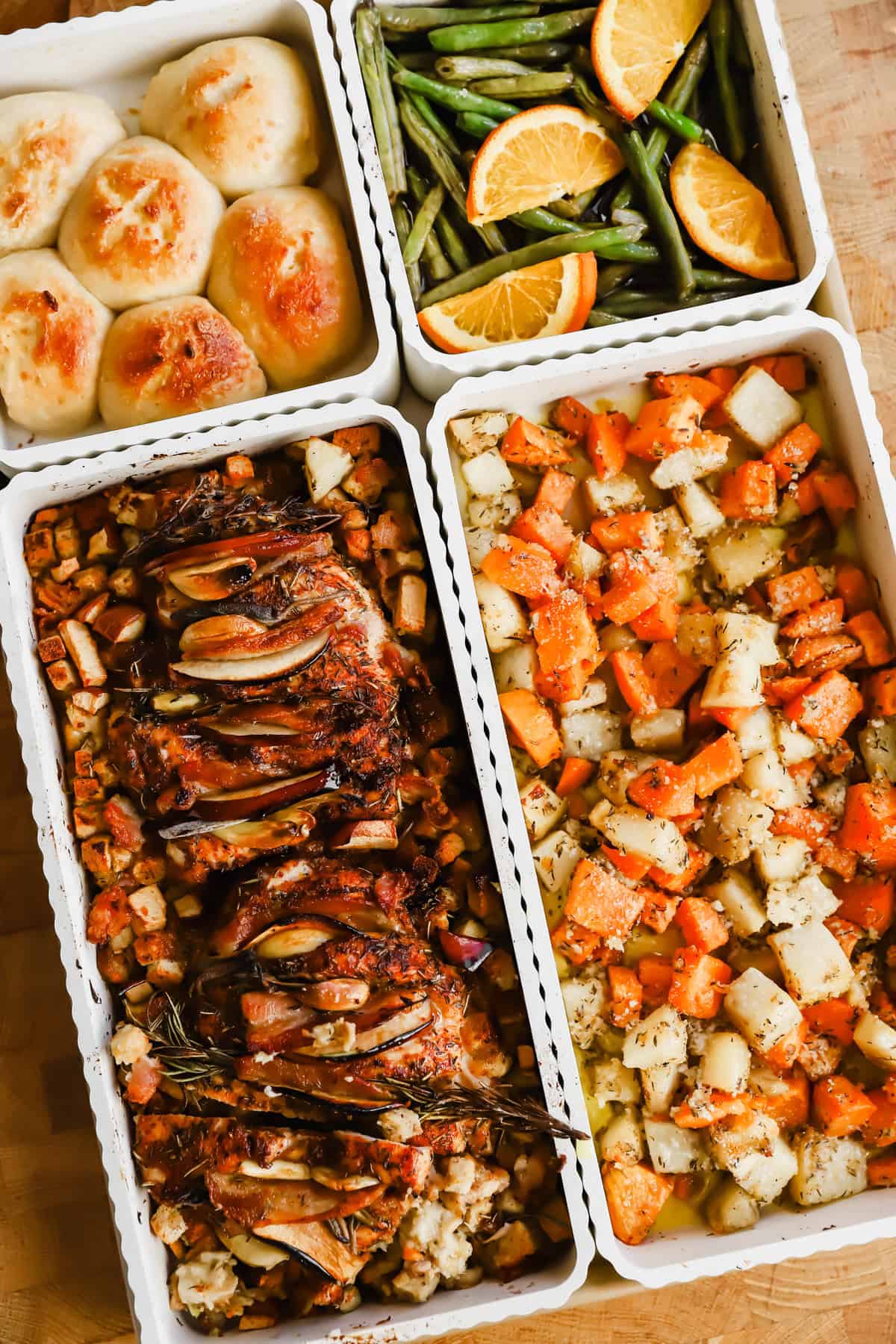 Four trays of food including dinner rolls, green beans with orange slices, roasted meat with herbs and vegetables, and a mix of roasted potatoes and carrots.