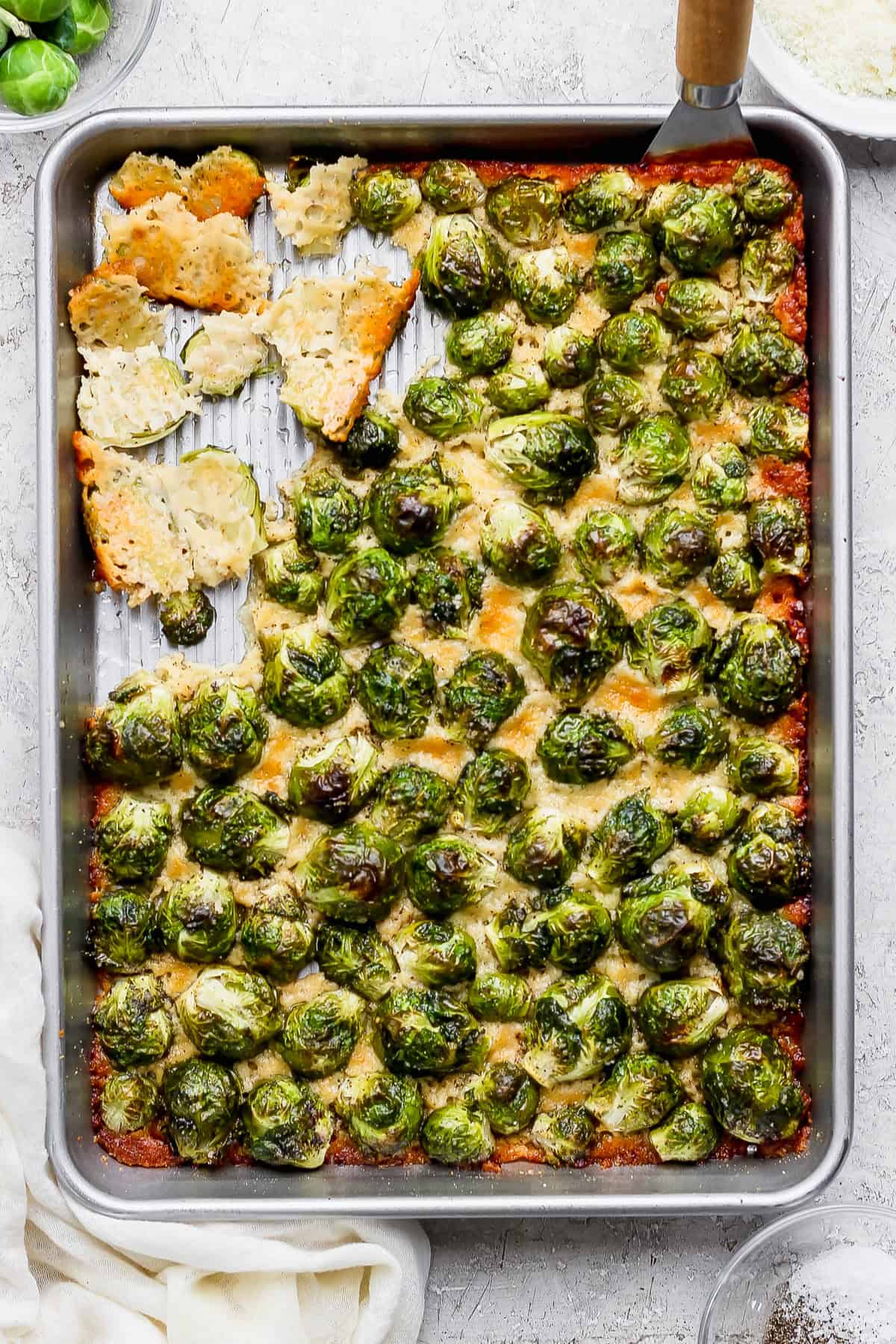 A baking sheet with roasted Brussels sprouts topped with melted cheese, partially cut into squares with a spatula, on a light-colored surface.