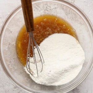 A glass bowl containing flour and a brown liquid being mixed with a metal whisk on a light textured surface.