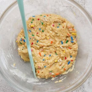 A glass bowl containing raw cookie dough with colorful sprinkles and a light blue spatula on a textured surface.