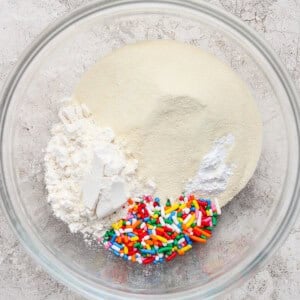 A glass bowl containing white flour, granulated sugar, baking powder, and colorful rainbow sprinkles on a light countertop.