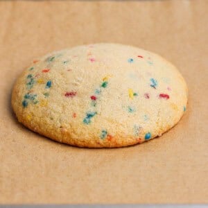 A single large sugar cookie with colorful sprinkles sits on a piece of parchment paper on a baking sheet.