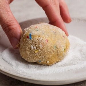 A hand rolls a ball of cookie dough with colorful sprinkles in a layer of white sugar on a plate.