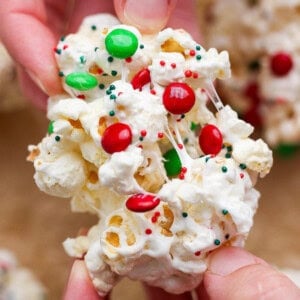 Close-up of hands holding a cluster of popcorn mixed with melted marshmallow, red and green candy-coated chocolates, and festive sprinkles.