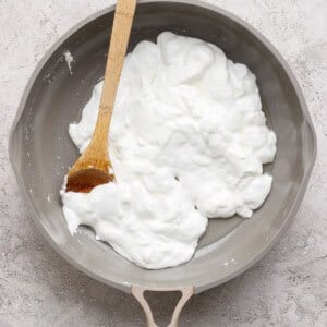 A nonstick skillet with a wooden spatula stirring a mound of whipped egg whites on a light textured surface.