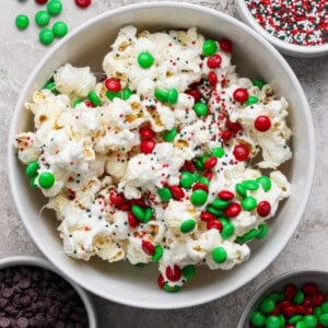A bowl of popcorn topped with red and green candy-coated chocolates and sprinkles, surrounded by bowls of chocolate chips, more candies, and sprinkles on a light surface.