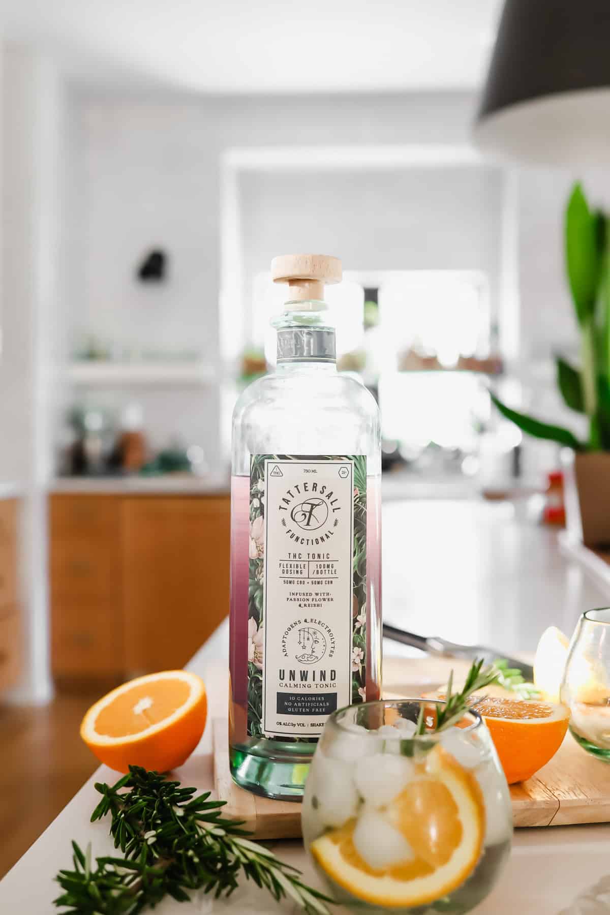 A bottle of Butterfly Spirits Botanical Flavored Gin on a kitchen counter, surrounded by sliced oranges, rosemary, and a glass of gin cocktail with ice and orange slices.