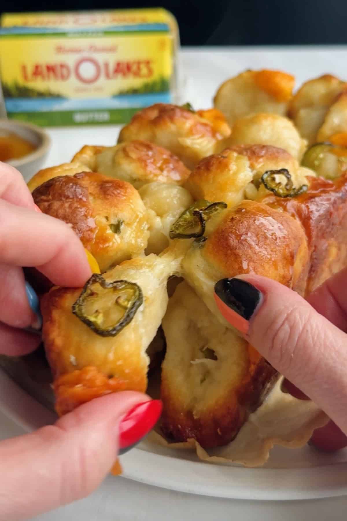 Two hands pull apart a piece of baked bread topped with sliced jalapeños, with a package of butter visible in the background.