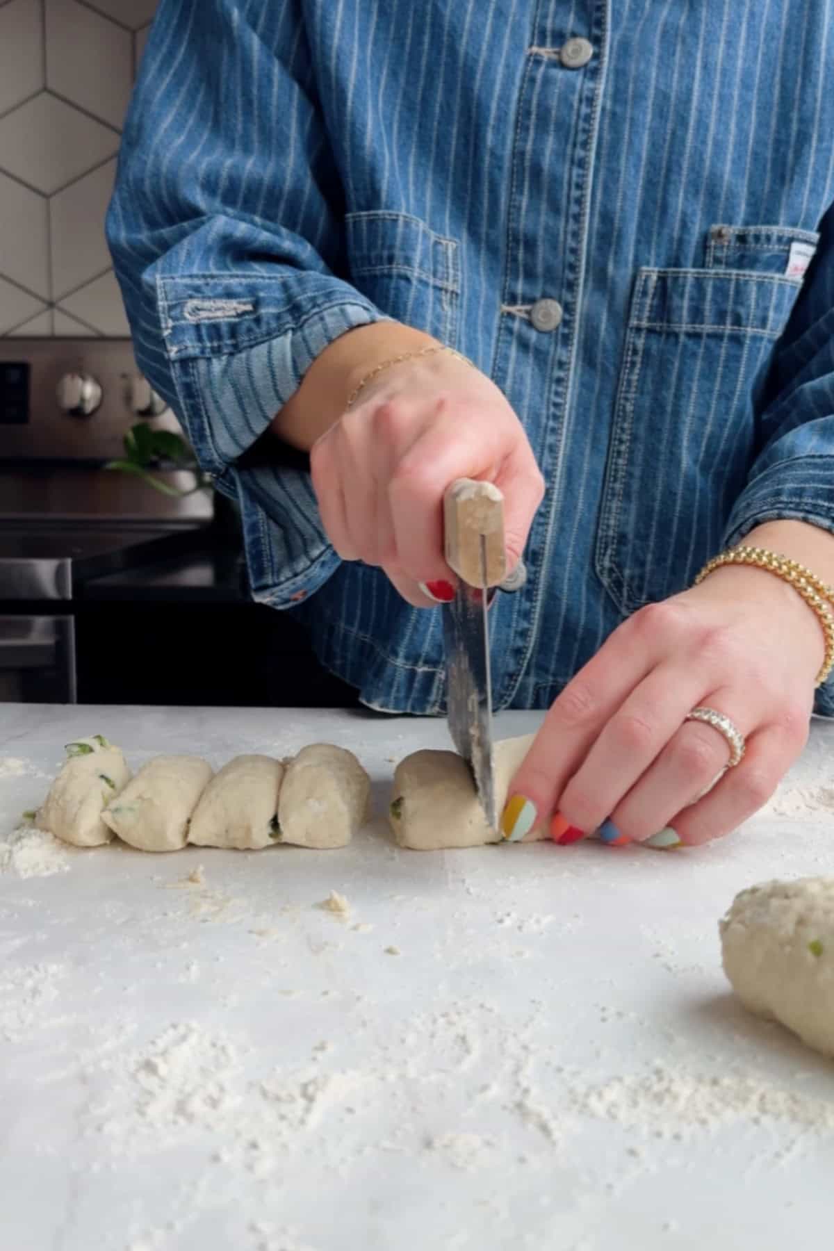A person in a denim shirt cuts a roll of dough into pieces on a floured surface in a kitchen.