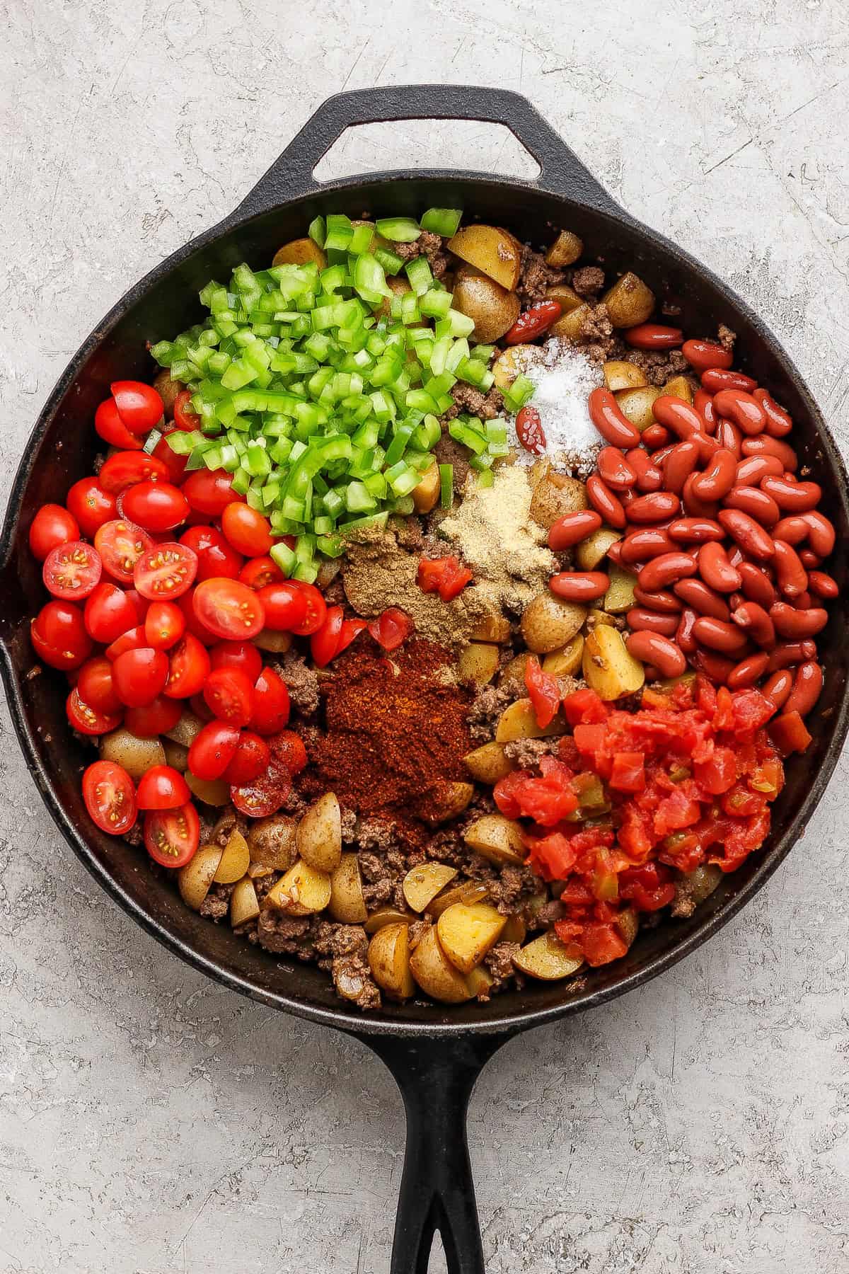 A cast iron skillet filled with diced potatoes, chopped tomatoes, kidney beans, green bell peppers, grape tomatoes, spices, and ground meat, all arranged in sections before mixing.