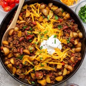 A skillet filled with diced potatoes, ground beef, kidney beans, and tomatoes, topped with shredded cheddar cheese, sour cream, and chopped parsley. A wooden spoon rests on the side.