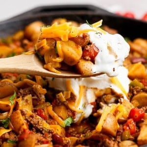 A wooden spoon holds a serving of skillet hash with ground beef, potatoes, vegetables, melted cheese, and sour cream over a cast iron pan filled with the same dish.