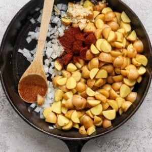 A cast iron skillet filled with chopped potatoes, diced onions, minced garlic, and paprika, with a wooden spoon resting on the side.