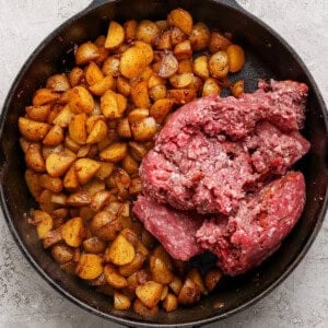 A cast iron skillet with cooked diced potatoes on one side and raw ground beef on the other, on a light textured surface.