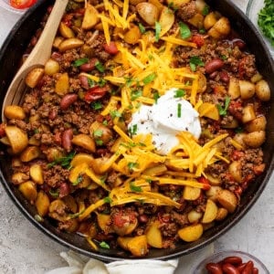 A skillet filled with ground beef, potatoes, kidney beans, and diced tomatoes, topped with shredded cheddar cheese and sour cream, surrounded by bowls of toppings.