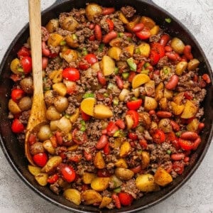 A cast iron skillet filled with cooked ground beef, halved baby potatoes, kidney beans, cherry tomatoes, and chopped green onions, with a wooden spoon resting inside.