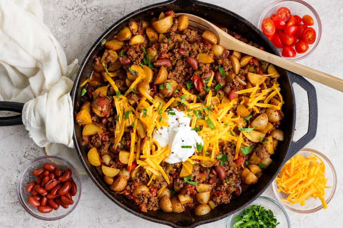 A cast iron skillet filled with chili, potatoes, kidney beans, ground meat, topped with shredded cheese, sour cream, and herbs. Bowls of tomatoes, beans, cheese, and herbs surround the skillet.