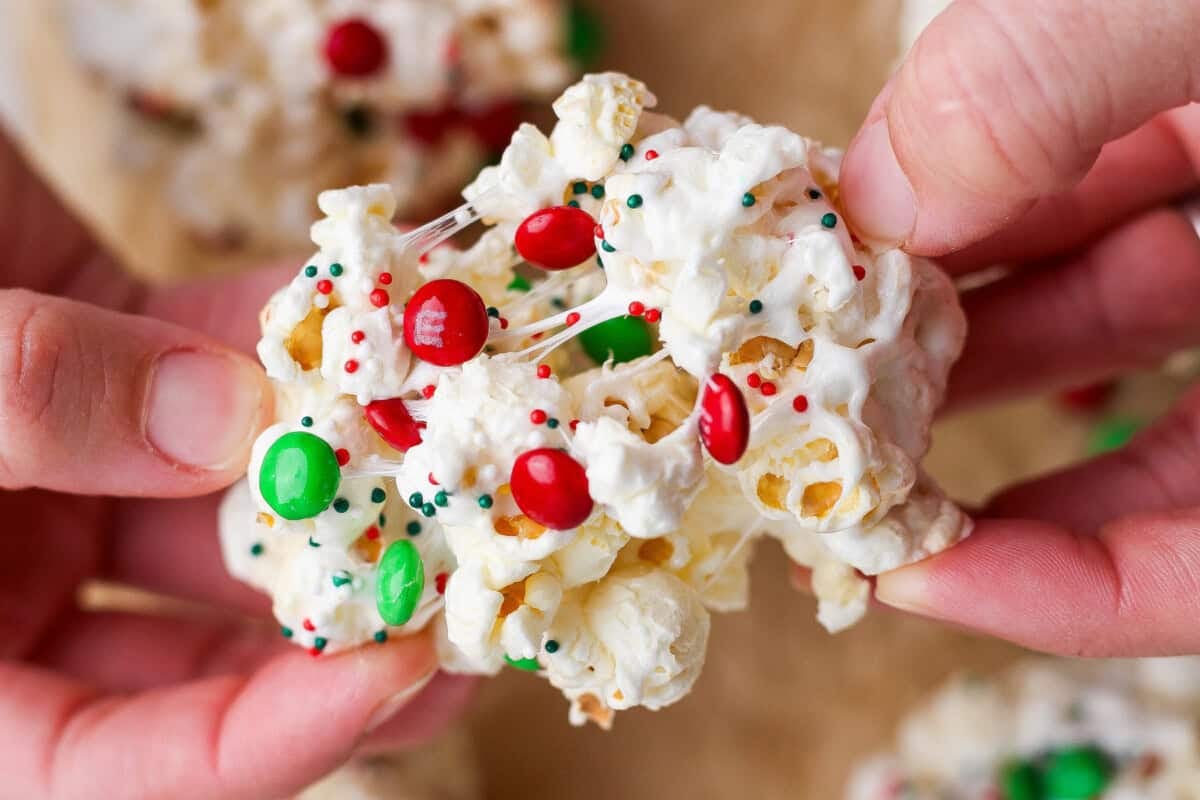 Close-up of hands holding a popcorn ball decorated with red and green candy-coated chocolates and festive sprinkles.