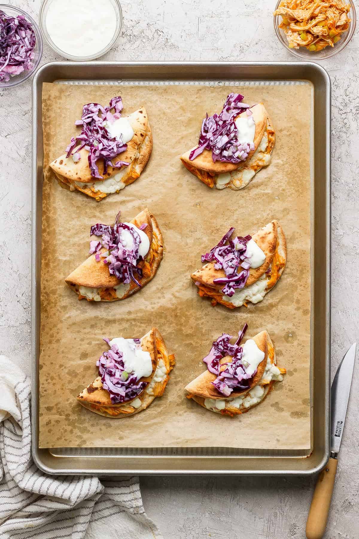 Six folded pita tacos filled with shredded meat, purple cabbage slaw, and a dollop of white sauce arranged on a parchment-lined baking sheet.