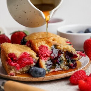 A white pitcher pours syrup over sliced berry-filled pancakes on a plate, surrounded by fresh strawberries, blueberries, and raspberries.