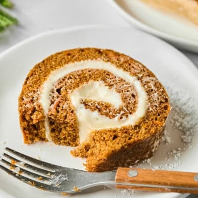 A slice of pumpkin roll with cream cheese filling on a white plate, partially eaten, with a fork beside it.
