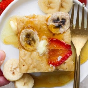 A square pancake topped with banana slices, a strawberry slice, melted butter, and syrup on a white plate with a gold fork and extra banana slices.