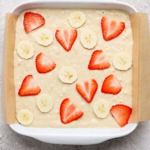 A baking dish lined with parchment paper contains unbaked batter topped with sliced strawberries and bananas.
