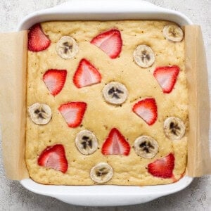 A square baking dish with a baked cake topped with sliced strawberries and bananas, lined with parchment paper on a light textured surface.
