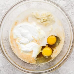 A glass bowl containing two raw eggs, yogurt, and other wet ingredients on a light textured surface.