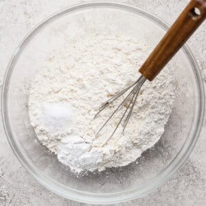 A glass bowl filled with flour and a small amount of salt, with a metal whisk that has a wooden handle resting inside.