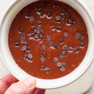 A hand holds a white bowl filled with chocolate batter topped with scattered chocolate chips.