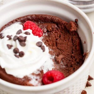 A chocolate mug cake topped with whipped cream, chocolate chips, and raspberries, with a portion eaten, in a white ramekin.