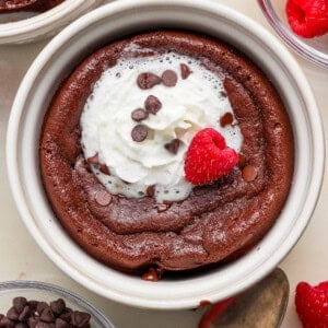 A chocolate lava cake in a white ramekin topped with whipped cream, chocolate chips, and a raspberry; surrounded by raspberries and chocolate chips.