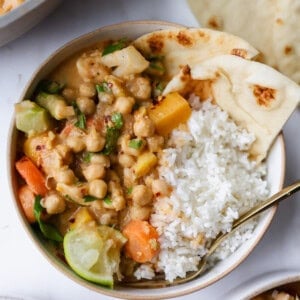 A bowl of chickpea curry with vegetables, white rice, lime wedge, and naan bread, served with a spoon on a white surface.