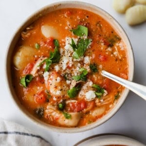 A bowl of tomato-based soup with gnocchi, shredded chicken, herbs, and cheese, with a spoon in the bowl and several gnocchi pieces and a napkin nearby.