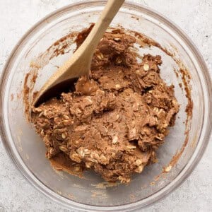 A glass bowl contains thick chocolate cookie dough with oats, being mixed with a wooden spoon, on a light textured surface.