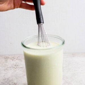 A hand holding a whisk over a glass jar filled with a creamy, pale liquid on a light-colored surface.