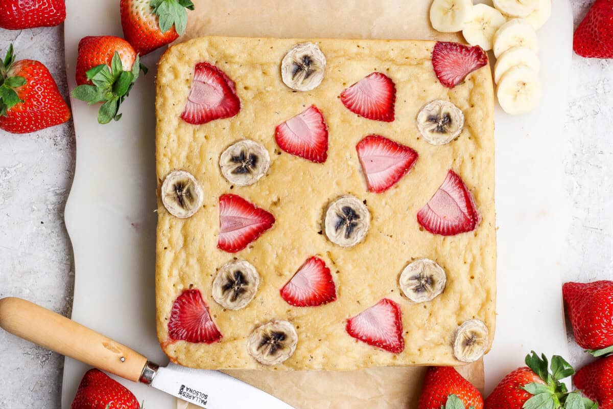 A square baked banana bread topped with sliced strawberries and bananas, surrounded by fresh strawberries and a knife on parchment paper.